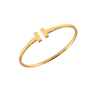 May include: A polished gold-tone open cuff bracelet featuring a sleek, circular band that terminates in two T-shaped ends facing each other. The metal has a smooth, reflective surface with a warm yellow hue, creating a continuous, minimalist silhouette.