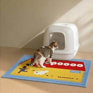 May include: A cat standing near a white enclosed litter box on a colorful mat. The mat has a yellow center with red and blue borders, and the text "pooooo" and "o!cute". The brand name "o!cute" is in blue.