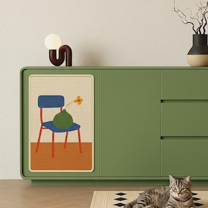 May include: A green cabinet with a decorative panel featuring a chair, vase, and flower illustration. A brown and white lamp sits on top. A tabby cat rests on a patterned rug in front of the cabinet.