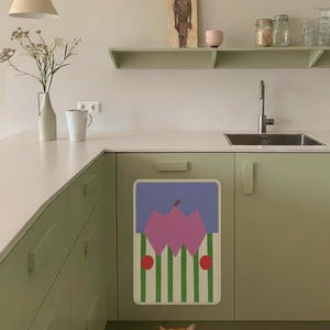 May include: A kitchen scene with a light green cabinet and a colorful abstract art piece on the cabinet door. A ginger cat is resting on the floor. A white vase with flowers and a white mug are on the countertop. A painting and shelves are on the wall.