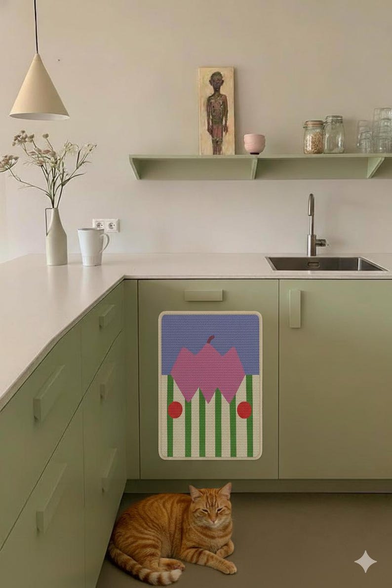 May include: A kitchen scene with a light green cabinet and a colorful abstract art piece on the cabinet door. A ginger cat is resting on the floor. A white vase with flowers and a white mug are on the countertop. A painting and shelves are on the wall.