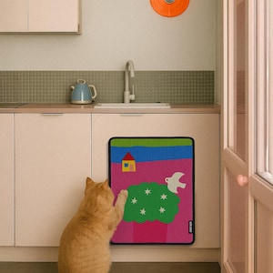 May include: A colorful, rectangular rug with a whimsical design featuring a house, a dove, and a green bush with stars. The rug is placed in front of a kitchen cabinet. An orange cat is looking at the rug.