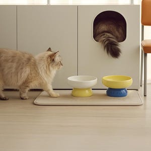 May include: A fluffy, light-colored cat walks past two ceramic pet bowls, one white and yellow, the other yellow and blue. A cat bed is built into a white cabinet, with a cat's tail visible. A beige mat sits beneath the bowls.