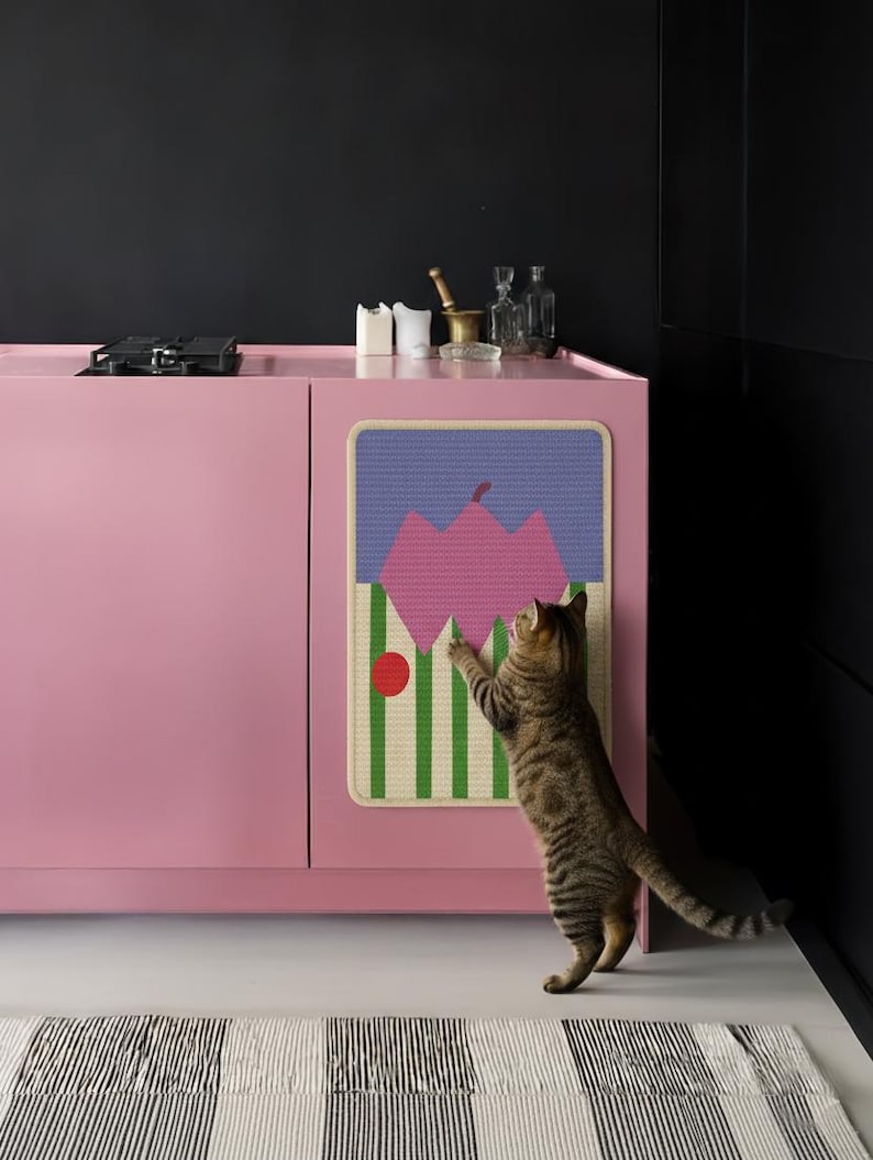 May include: A pink cabinet with a colorful, geometric cat scratcher attached. A tabby cat stands on its hind legs, reaching towards the scratcher. A striped rug is in the foreground.