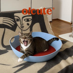 May include: A yawning tabby cat rests in a light blue pet bed with a red cushion. The bed has handles and is on a patterned blanket. The word "o!cute" is in orange letters above the cat.