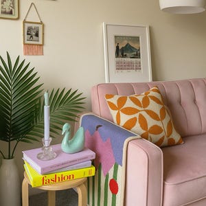 May include: A cozy living room scene with a pink sofa, patterned throw blanket, and decorative pillows. A small wooden side table holds books, a swan figurine, and a candle. Framed art and a palm plant add to the decor.