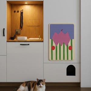 May include: A calico cat rests on a dark wood floor in front of a white cabinet with a cat door. A colorful wall hanging with a pink mountain design and green and white stripes hangs above the cat door. The cabinet has a built-in shelf with a light.