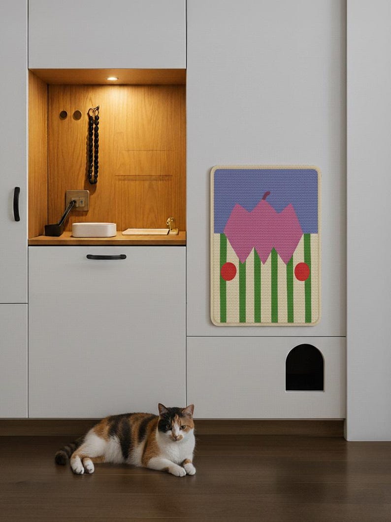 May include: A calico cat rests on a dark wood floor in front of a white cabinet with a cat door. A colorful wall hanging with a pink mountain design and green and white stripes hangs above the cat door. The cabinet has a built-in shelf with a light.