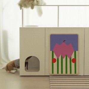 May include: A modern cat furniture set with a beige cabinet, a decorative mat with a geometric design, and a fluffy cat resting on top. The mat features a pink and green pattern. A dog is partially visible inside the cabinet.