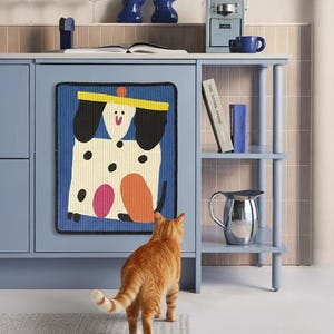 May include: A whimsical, colorful dog-themed wall hanging is mounted on a blue cabinet. The image also features a blue coffee maker, a blue sculpture, and a silver pitcher. An orange cat stands in front of the cabinet, looking at the wall hanging.