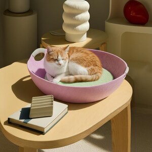 May include: A ginger and white cat rests in a pink cat bed with a light green cushion. The bed is on a light wood table. A white lamp with a ribbed design and a small book are also on the table. The scene is brightly lit.