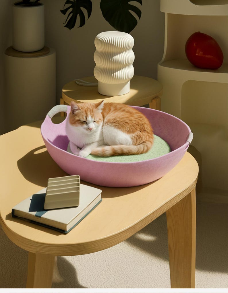 May include: A ginger and white cat rests in a pink cat bed with a light green cushion. The bed is on a light wood table. A white lamp with a ribbed design and a small book are also on the table. The scene is brightly lit.