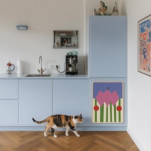 May include: A calico cat walks across a herringbone wood floor in a kitchen with light blue cabinets. A framed artwork with pink and green shapes hangs on the cabinet. A coffee maker and a framed photo are on the counter.