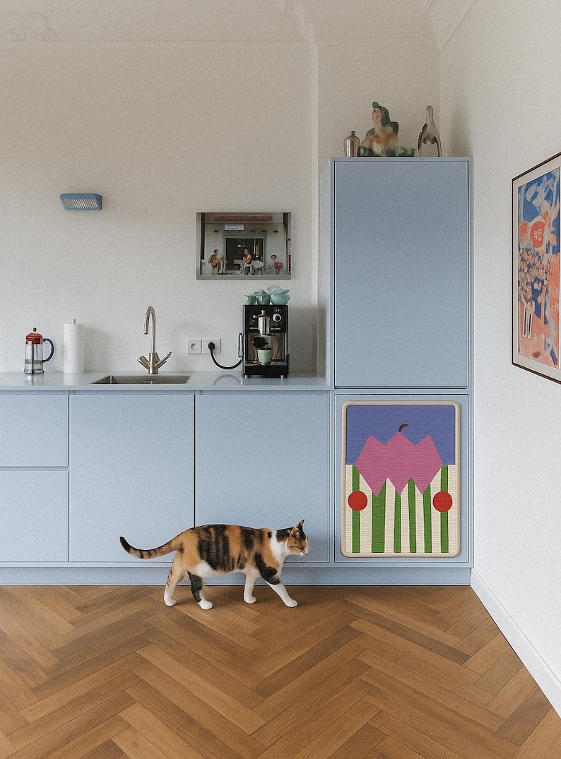 May include: A calico cat walks across a herringbone wood floor in a kitchen with light blue cabinets. A framed artwork with pink and green shapes hangs on the cabinet. A coffee maker and a framed photo are on the counter.