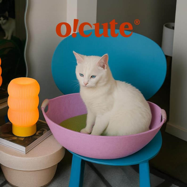 May include: A white cat with blue eyes sits in a pink pet bed on a blue chair. The bed has a green cushion. An orange lamp and the text "o!cute" are also visible.