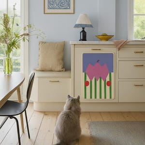 May include: A kitchen scene with a light blue wall, a beige cabinet, and a decorative panel featuring a pink floral design with green stripes and red dots. A gray cat sits on the wooden floor, looking at the panel. A beige cushion sits on a bench.