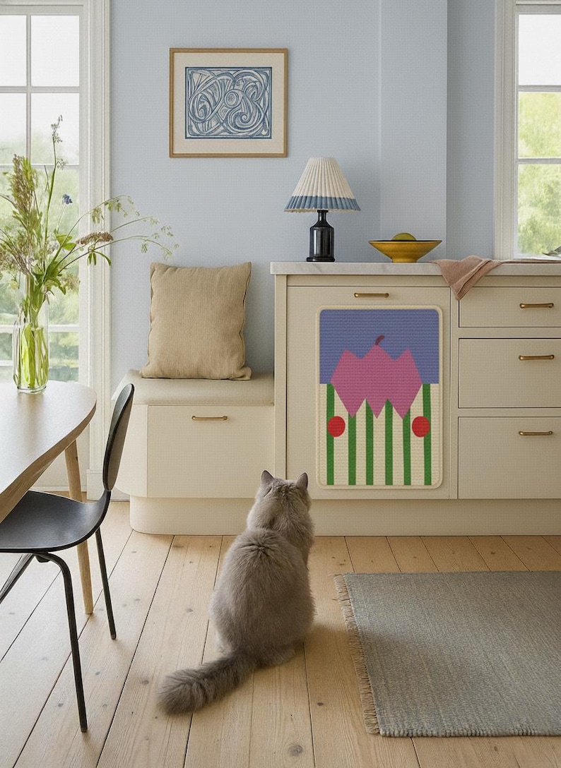 May include: A kitchen scene with a light blue wall, a beige cabinet, and a decorative panel featuring a pink floral design with green stripes and red dots. A gray cat sits on the wooden floor, looking at the panel. A beige cushion sits on a bench.