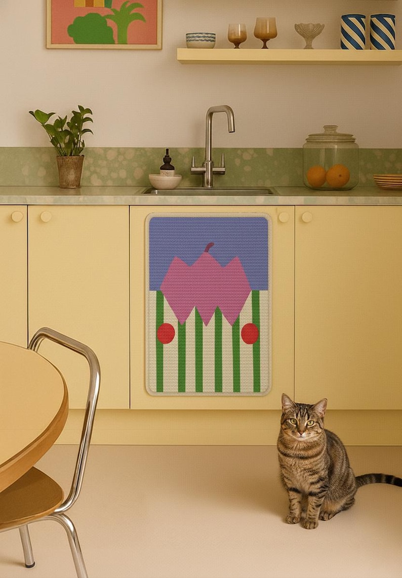 May include: A kitchen scene featuring a decorative mat with a pink and green design, set against yellow cabinets and a light green countertop. A tabby cat sits in the foreground.