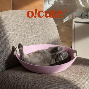 May include: A gray cat is comfortably resting in a pink felt cat bed. The bed has a curved design with white handles. The image includes the text "o!cute" in orange. The cat is lying on its back on a gray upholstered chair.