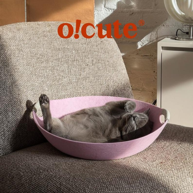 May include: A gray cat is comfortably resting in a pink felt cat bed. The bed has a curved design with white handles. The image includes the text "o!cute" in orange. The cat is lying on its back on a gray upholstered chair.