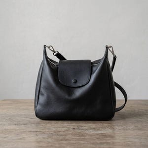 May include: A black leather shoulder bag with a flap and button closure. The bag features a zip closure and an adjustable strap. The bag is on a wooden surface.
