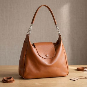 May include: A tan leather shoulder bag with a curved handle and silver-tone hardware. The bag features a flap closure with a single button. A small leather strap and a tag are visible.