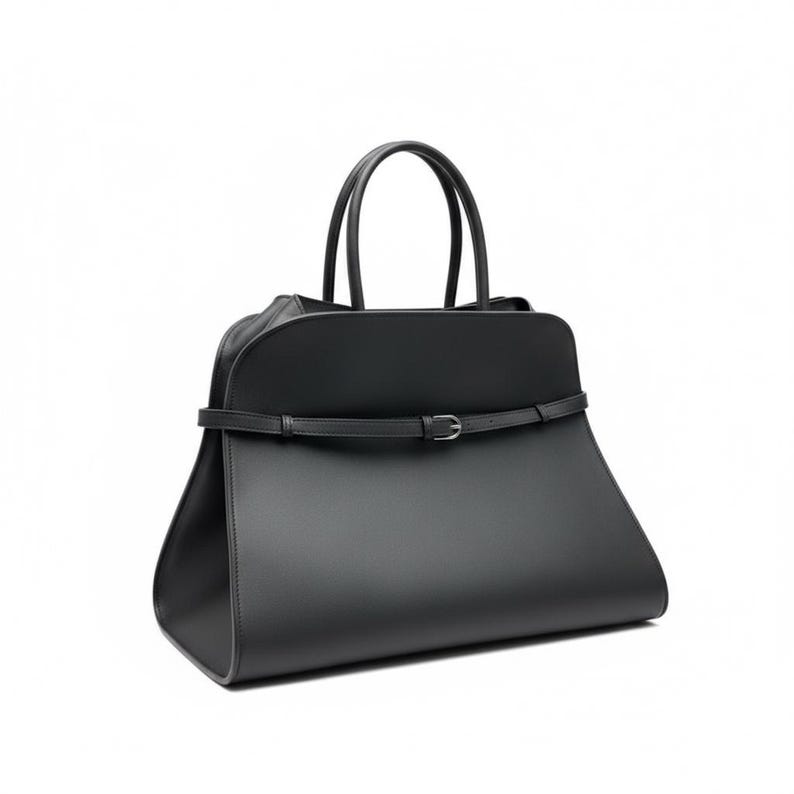 May include: A black handbag with a trapezoid shape and two top handles. The bag features a thin belt with a silver buckle around the middle. The bag is made of a smooth material and has a minimalist design.