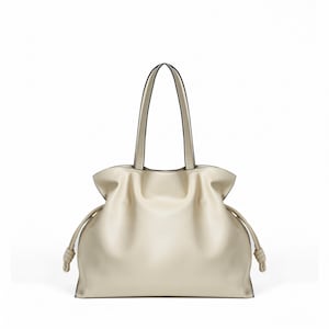 May include: A cream-colored tote bag with a soft, slouchy silhouette, featuring two thin shoulder straps and side drawstrings that end in knotted details. The bag has a smooth, matte finish and a wide, open-top design with gathered sides.