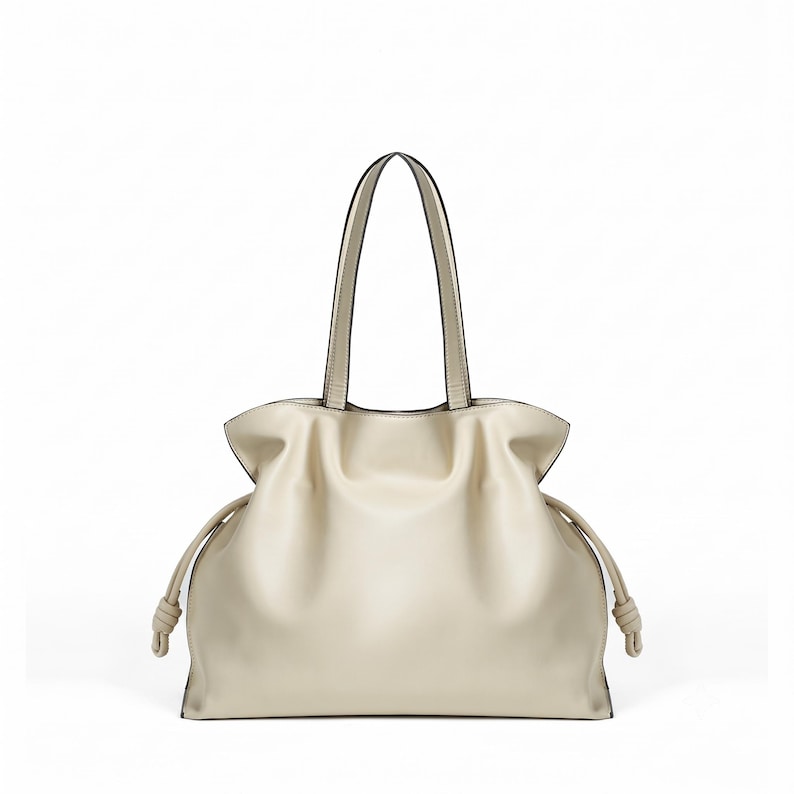 May include: A cream-colored tote bag with a soft, slouchy silhouette, featuring two thin shoulder straps and side drawstrings that end in knotted details. The bag has a smooth, matte finish and a wide, open-top design with gathered sides.
