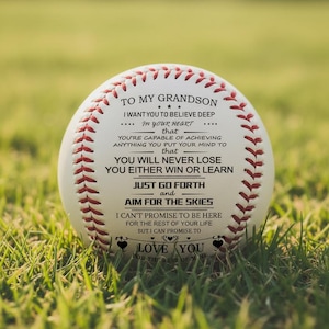 May include: A white baseball with red stitching and text. The text reads "TO MY GRANDSON" and includes an inspirational message. The baseball is resting in green grass, suggesting an outdoor setting. A heartfelt gift for a grandson.
