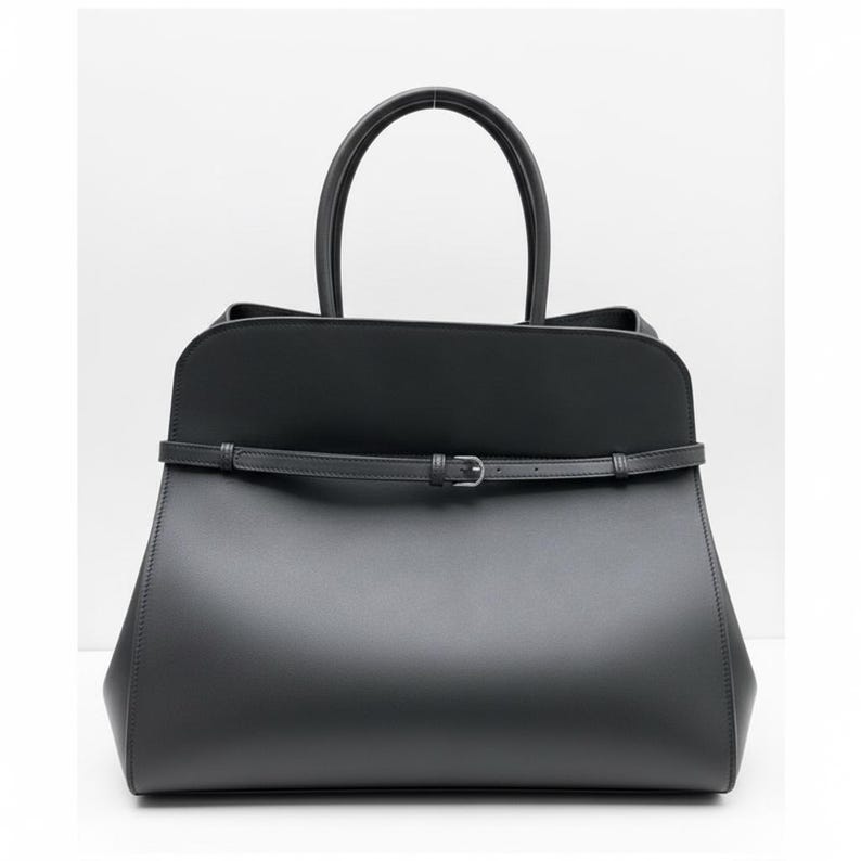 May include: A black tote bag with a structured design. The bag has a top handle and a thin strap with a buckle. The bag's shape is trapezoidal, with a wider base and a narrower top.