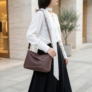 Puede incluir: Bolso de hombro de cuero marrón con cierre de solapa y costuras blancas. El bolso tiene una correa larga y fina y herrajes dorados. Una persona lleva el bolso, vestida con una blusa blanca y una falda negra.