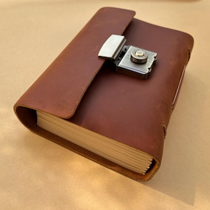 Könnte beinhalten: Ein braunes Lederjournal mit einem Metallverschluss. Das Journal hat einen Klappenverschluss und einen Verriegelungsmechanismus. Die Seiten sind am Rand sichtbar. Das Leder hat ein abgenutztes Aussehen. Das Journal ist geschlossen und liegt auf einer hellbraunen Oberfläche.