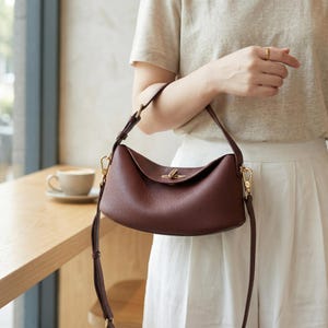 Puede incluir: Un bolso de mano de cuero burdeos con forma curva y asa superior. El bolso tiene un cierre y herrajes dorados, y una correa larga y ajustable. El bolso lo sostiene una persona que lleva una camisa clara y pantalones blancos.