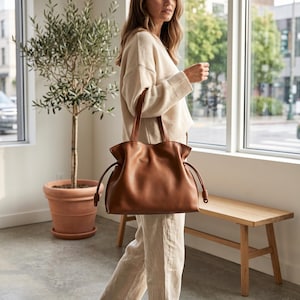 May include: A smooth, cognac-colored leather tote bag featuring a soft, slouchy silhouette, two sturdy shoulder straps, and side drawstring closures with knotted ends that create a gathered, ruffled effect along the top opening of the bag.