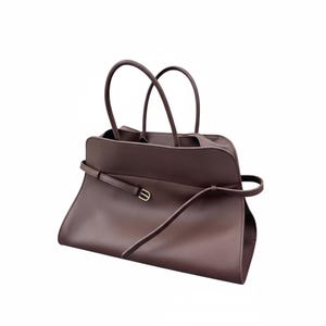 May include: A brown leather tote bag with two curved handles and a long, thin strap that wraps around the body. The bag has a rectangular shape with a subtle curve at the top and a small gold-coloured buckle.