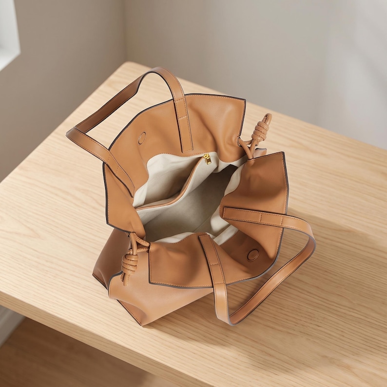 May include: A tan tote bag with a smooth finish, featuring two top handles and knotted side details. The interior is lined with a light-colored fabric and includes a central divider pocket with a gold-toned zipper pull, providing an open-top design.