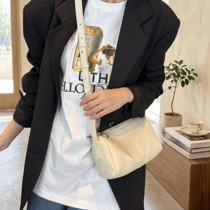 Puede incluir: Bolso de hombro color crema con un panel superior gris claro. El bolso tiene cierre de cremallera y una correa larga y ajustable. La persona lleva una camiseta blanca con un estampado gráfico y una blazer negra.