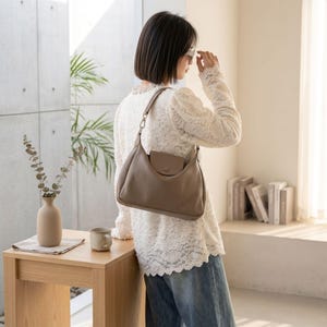 May include: A light brown shoulder bag with a small front pocket and a single strap. The bag is made of a soft material and has a rounded shape. The bag is being worn over the shoulder. The background includes a wooden table, a vase, and a mug.