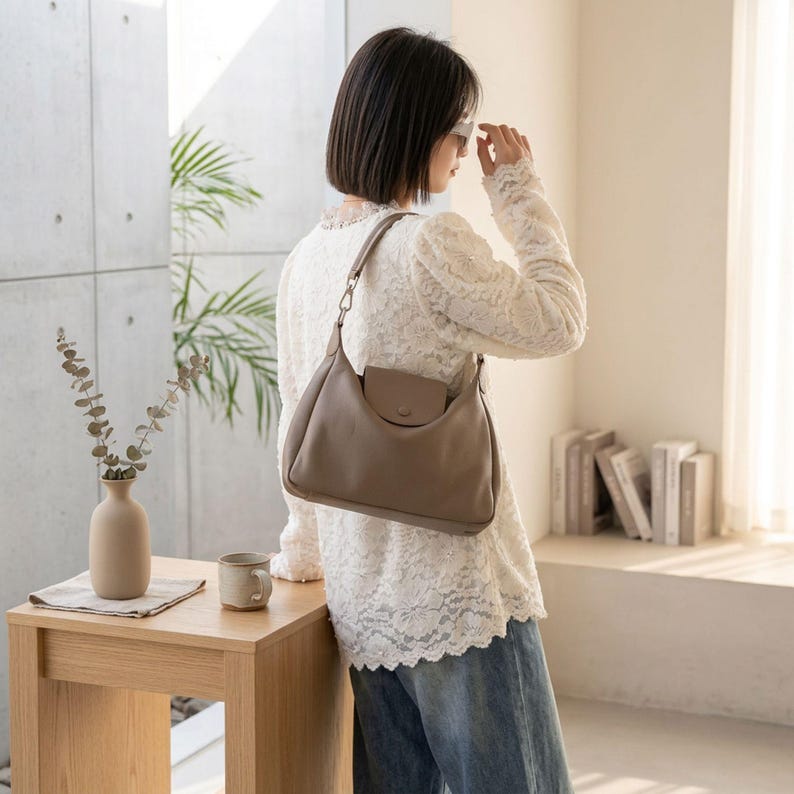 May include: A light brown shoulder bag with a small front pocket and a single strap. The bag is made of a soft material and has a rounded shape. The bag is being worn over the shoulder. The background includes a wooden table, a vase, and a mug.