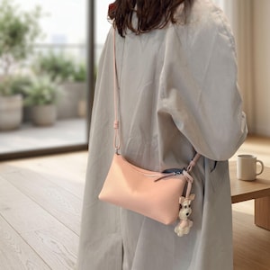 May include: A light pink, rectangular crossbody bag with a zipper closure and a matching strap. A small dog-shaped charm hangs from the strap. The bag is made of a textured material.