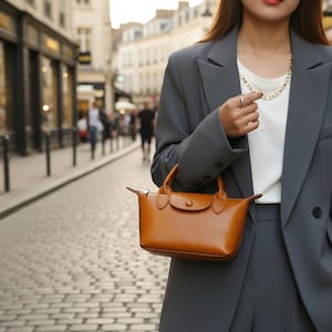 May include: A small, rectangular, brown leather handbag with a top handle and a flap closure. The bag is held by a person wearing a gray blazer and white shirt. The background shows a cobblestone street and buildings.