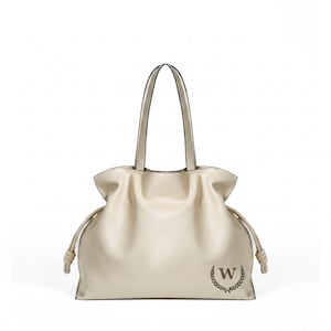 May include: A cream-colored tote bag with two shoulder straps, featuring a gathered top design with side drawstring cords ending in knots. The front lower right corner displays a dark brown letter W enclosed within a circular laurel wreath emblem.