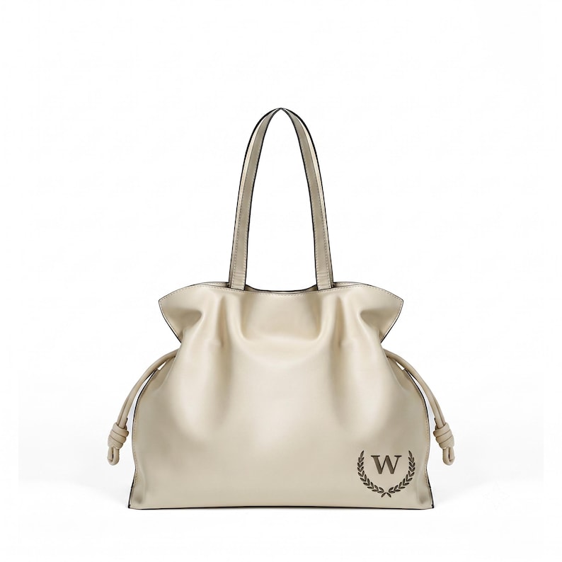 May include: A cream-colored tote bag with two shoulder straps, featuring a gathered top design with side drawstring cords ending in knots. The front lower right corner displays a dark brown letter W enclosed within a circular laurel wreath emblem.