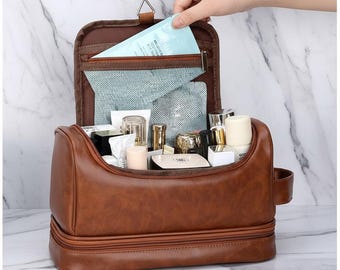 May include: A brown leather-effect hanging wash bag, open to show various cosmetic products. The bag has a top compartment with a zip and a mesh pocket. A hand is reaching into the top compartment. The bag is filled with bottles, tubes, and containers.