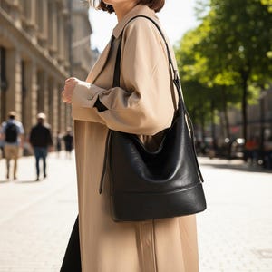 May include: A black leather shoulder bag with a rounded shape and a flat bottom. The bag has a long strap and is being carried by someone wearing a tan trench coat. The background shows a city street with people and trees.