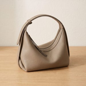 Könnte beinhalten: Eine taupefarbene Handtasche mit geschwungenem Design. Die Tasche hat einen oberen Griff und einen Reißverschluss. Die Ledertasche liegt auf einer Holzoberfläche. Die Tasche hat ein minimalistisches Design.