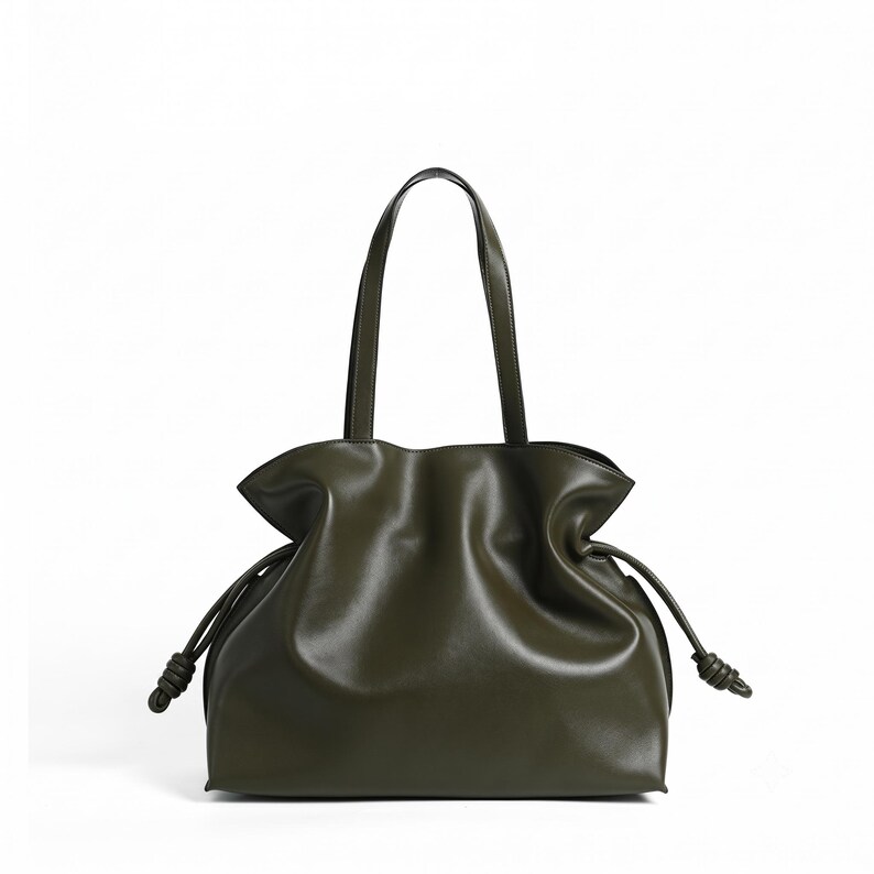 May include: An olive green shoulder bag with a smooth finish, featuring a gathered top opening, two thin shoulder straps, and side drawstrings that end in knotted details. The bag has a soft, slouchy structure with a flat base and curved sides.