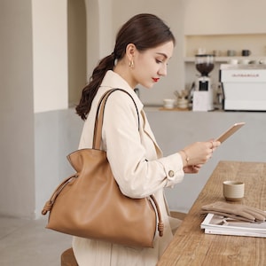 May include: A tan leather tote bag with a soft, slouchy silhouette, featuring thin shoulder straps and decorative knotted drawstring details on the sides. The bag has a smooth, matte finish and a wide, open-top design for easy access to contents.