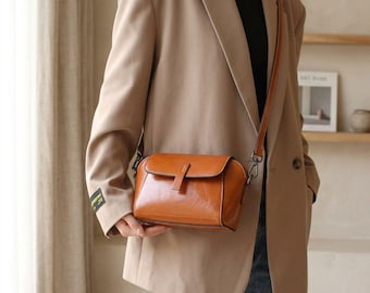 May include: A brown leather crossbody bag with a flap closure and a long, adjustable strap. The bag has a rectangular shape and a glossy finish. The hardware is silver-toned. The bag is held in front of a person wearing a tan blazer.
